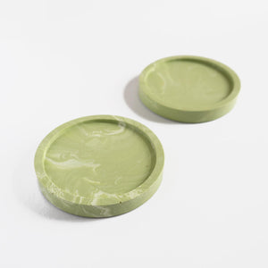 A pair of handmade round coasters with a sage green marbled pattern by Klndra for Curious Makers